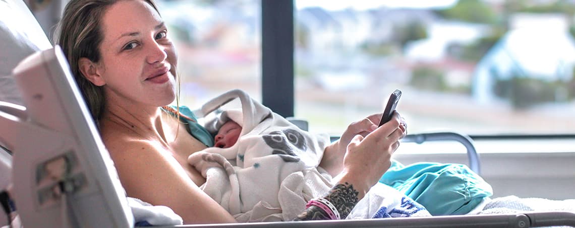 一个微笑的新妈妈和她的新生儿在医院的病床上打电话